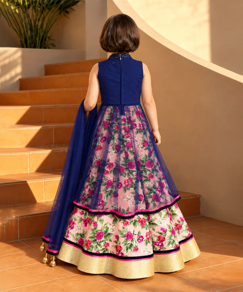 A young girl viewed from behind wearing a traditional Indian anarkali-style dress with a navy blue sleeveless bodice and a heavily embroidered pink floral skirt with a matching navy dupatta, standing on terracotta stairs.