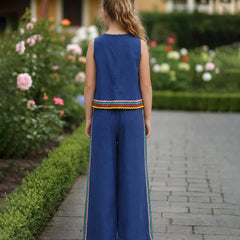 A young girl viewed from behind wearing a sleeveless royal blue jumpsuit with colorful striped trim at the hem and down the sides of the wide legs, standing on a stone path in a garden.