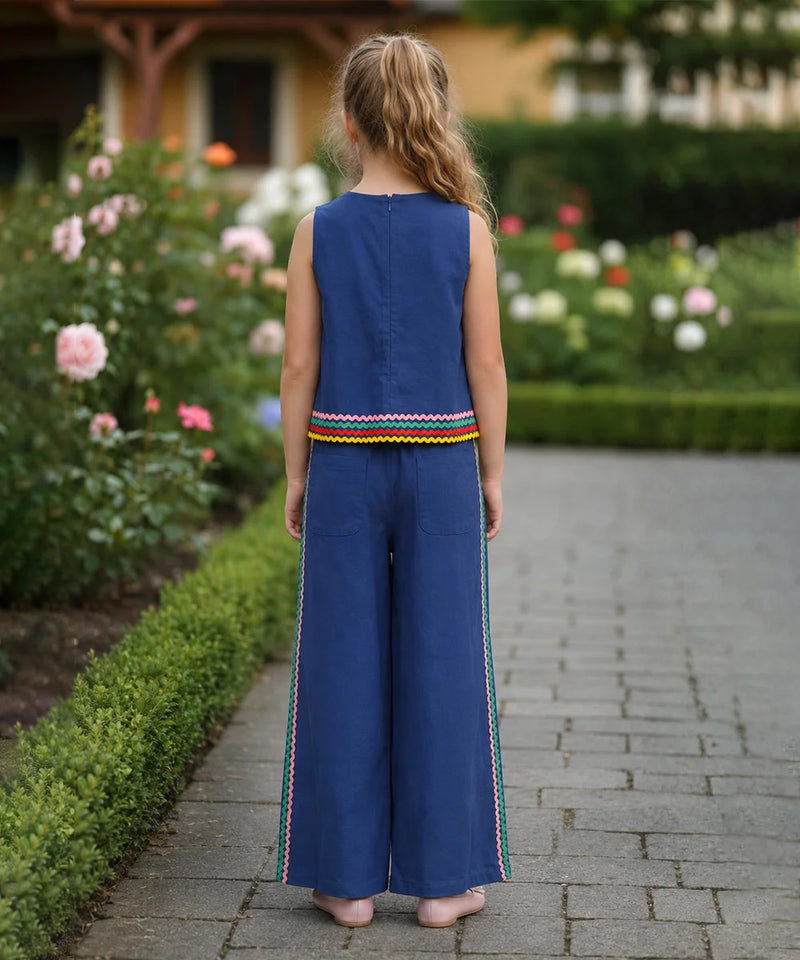 A young girl viewed from behind wearing a sleeveless royal blue jumpsuit with colorful striped trim at the hem and down the sides of the wide legs, standing on a stone path in a garden.