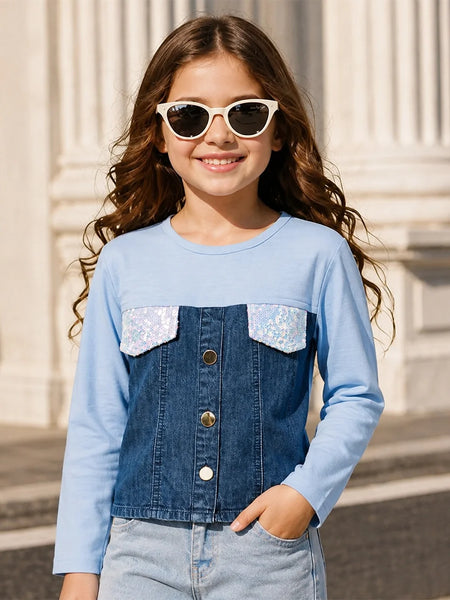 A young girl with long wavy brown hair and white cat-eye sunglasses is wearing a light blue long-sleeve top with a dark blue denim-style front panel and two silver sequined chest pockets. She is posing outdoors in front of a light-colored stone building.