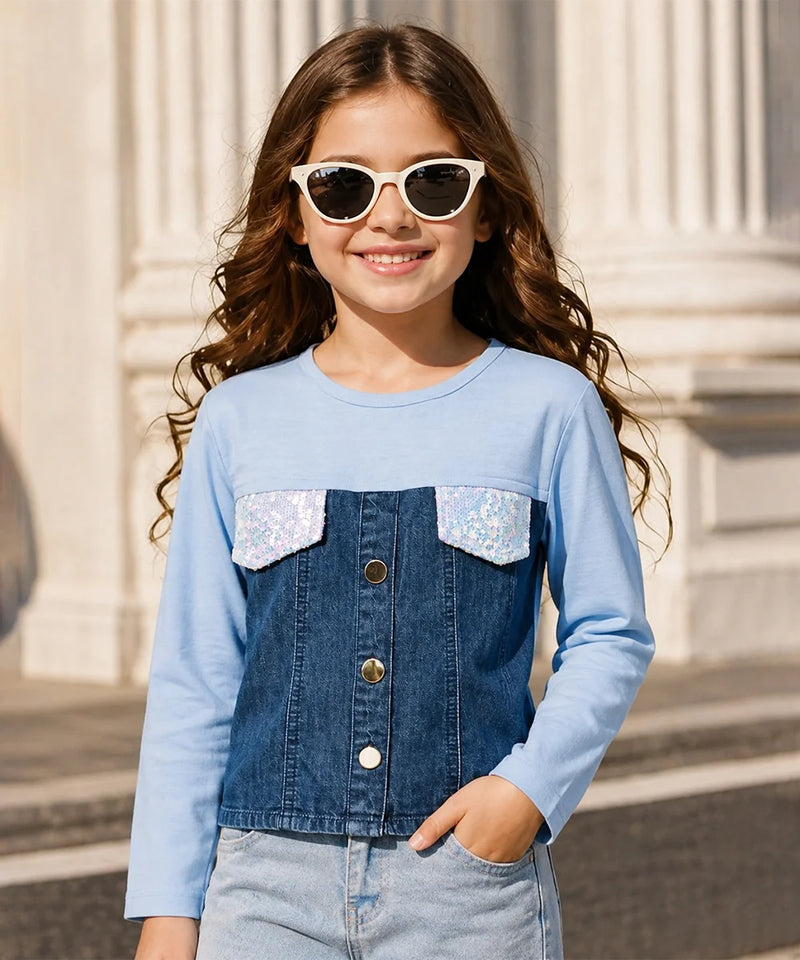 A young girl with long wavy brown hair and white cat-eye sunglasses is wearing a light blue long-sleeve top with a dark blue denim-style front panel and two silver sequined chest pockets. She is posing outdoors in front of a light-colored stone building.