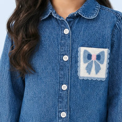 A close-up of a child wearing a blue denim button-up shirt with a decorative patch pocket featuring a blue bow and pink center on a white background.