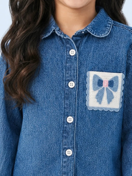 A close-up of a child wearing a blue denim button-up shirt with a decorative patch pocket featuring a blue bow and pink center on a white background.