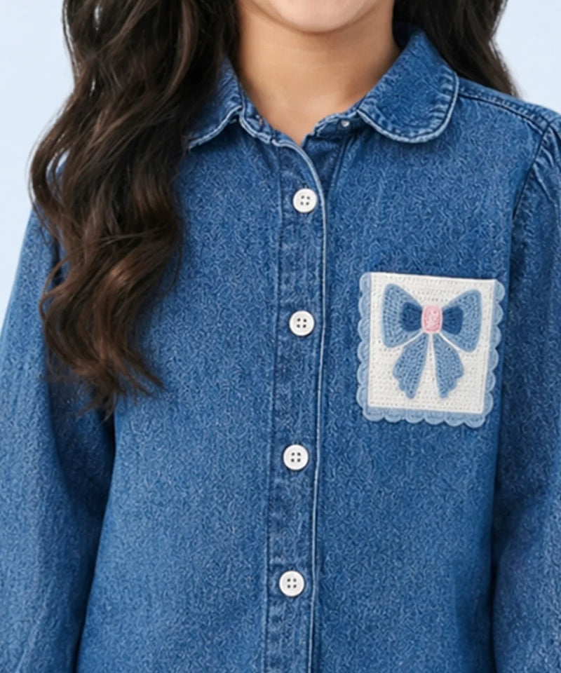 A close-up of a child wearing a blue denim button-up shirt with a decorative patch pocket featuring a blue bow and pink center on a white background.