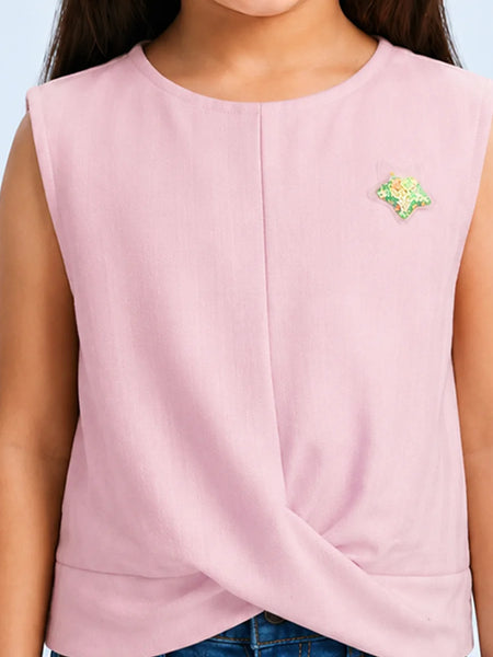 A sleeveless light pink top with a twisted hem detail, worn by a model. The top features a small green and yellow embroidered star on the left chest.