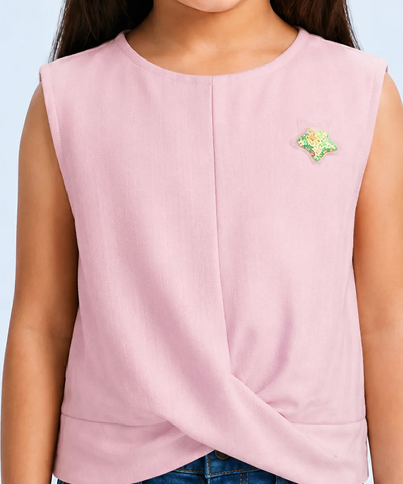 A sleeveless light pink top with a twisted hem detail, worn by a model. The top features a small green and yellow embroidered star on the left chest.