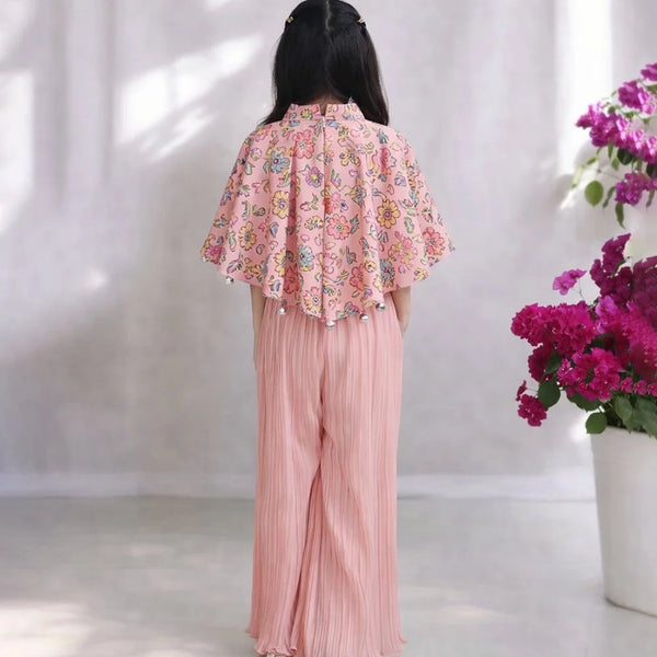 Person wearing a pink floral blouse and pleated pants with a white curtain and flowers in the background.