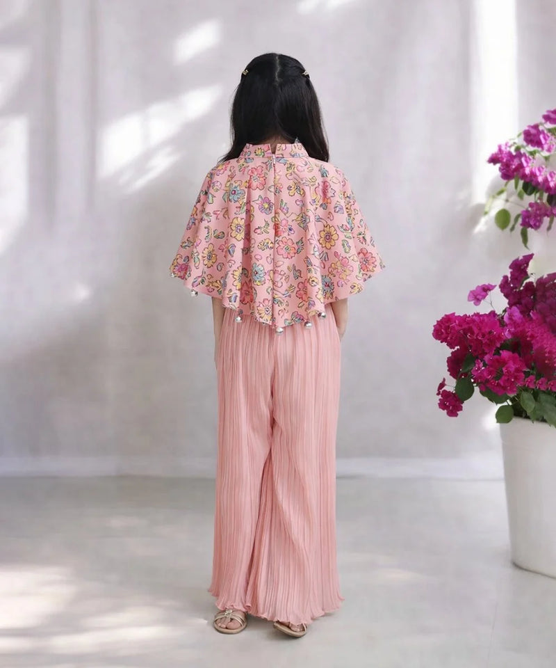 Person wearing a pink floral blouse and pleated pants with a white curtain and flowers in the background.