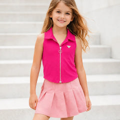 A young girl with long light brown hair stands on light gray concrete steps, wearing a sleeveless bright pink collared zip-front top with a small white geometric logo on the chest and a light pink pleated skirt.