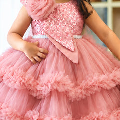 A young girl wearing a pink sequined and tulle party dress with a large floral embellishment on the shoulder and layered ruffle tiers.