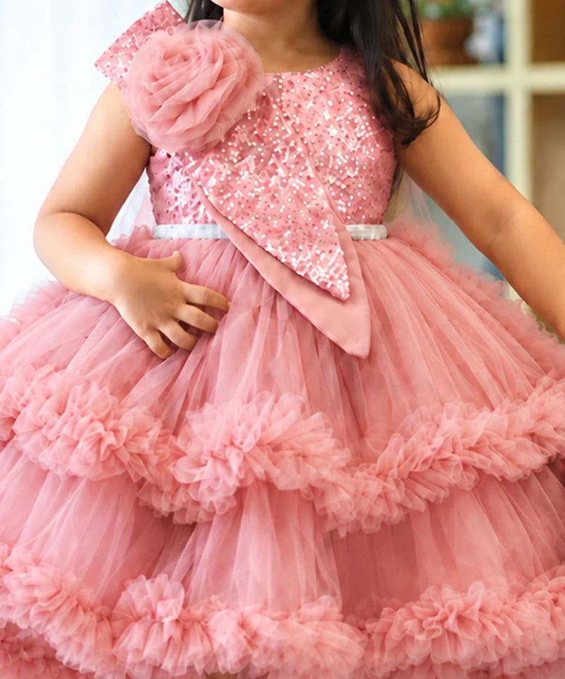 A young girl wearing a pink sequined and tulle party dress with a large floral embellishment on the shoulder and layered ruffle tiers.