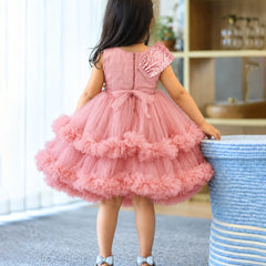 A young girl viewed from behind wearing a pink tulle dress with a ruffled tiered skirt and a single embellished sleeve, standing indoors next to a blue-and-white striped basket.
