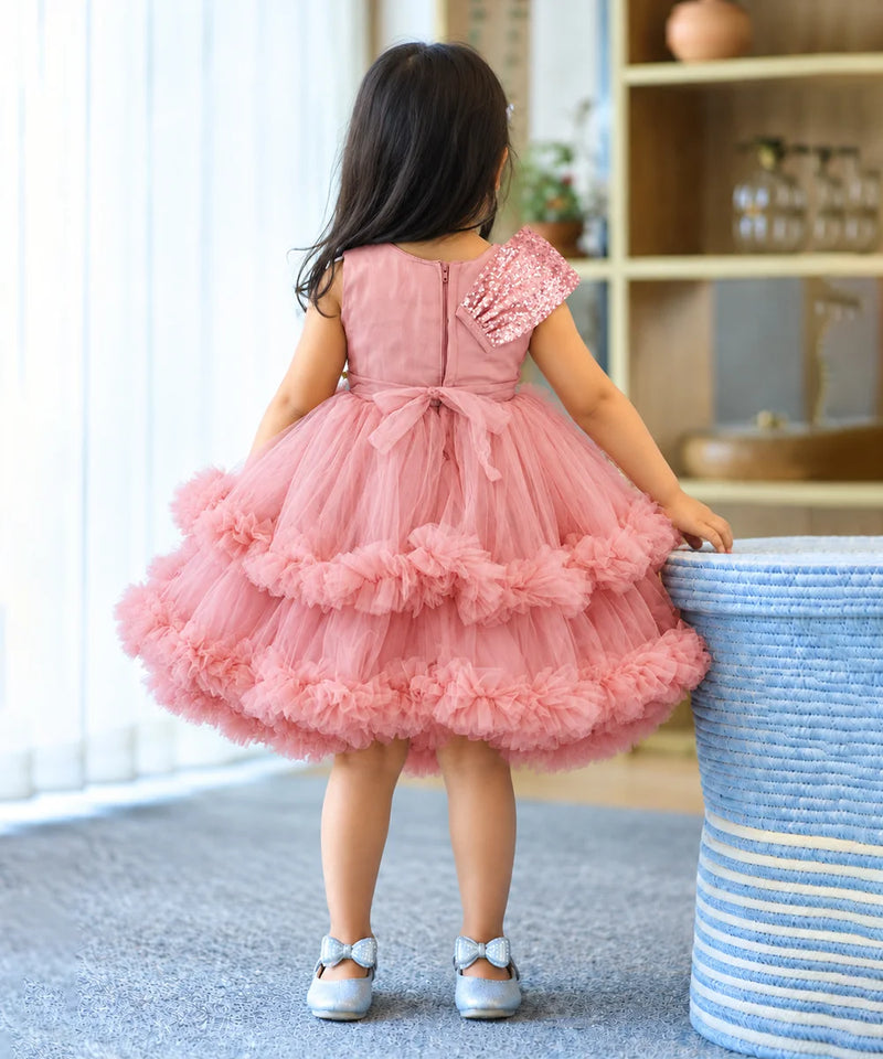A young girl viewed from behind wearing a pink tulle dress with a ruffled tiered skirt and a single embellished sleeve, standing indoors next to a blue-and-white striped basket.