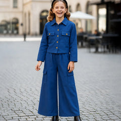 A young girl stands smiling in a cobblestone plaza wearing a matching blue two-piece outfit consisting of a cropped button-front jacket with chest pockets and wide-leg cargo pants, paired with black boots and a black headband.