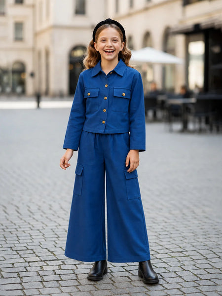 A young girl stands smiling in a cobblestone plaza wearing a matching blue two-piece outfit consisting of a cropped button-front jacket with chest pockets and wide-leg cargo pants, paired with black boots and a black headband.