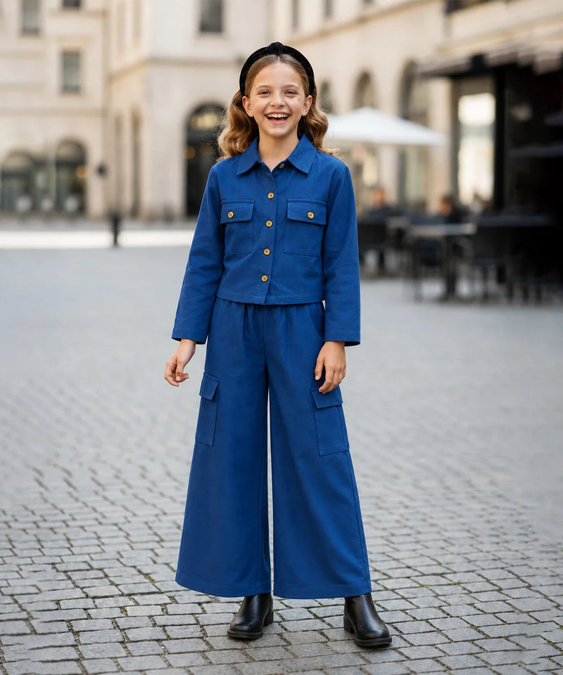 A young girl stands smiling in a cobblestone plaza wearing a matching blue two-piece outfit consisting of a cropped button-front jacket with chest pockets and wide-leg cargo pants, paired with black boots and a black headband.