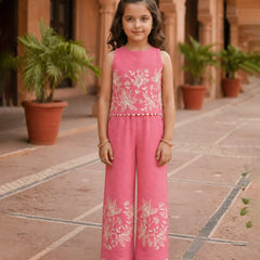 Young girl in a pink embroidered outfit standing in an outdoor setting with plants and architectural elements.