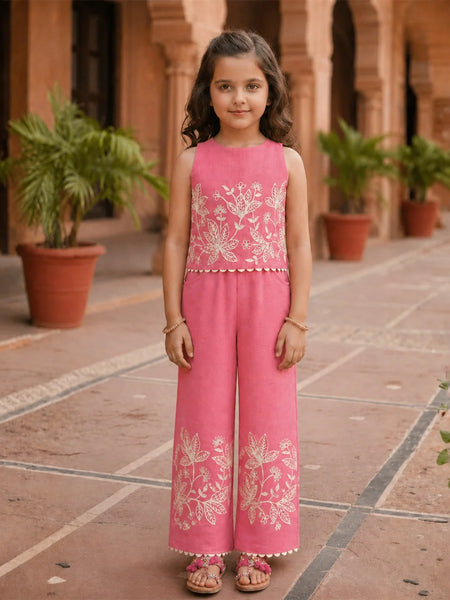 Young girl in a pink embroidered outfit standing in an outdoor setting with plants and architectural elements.