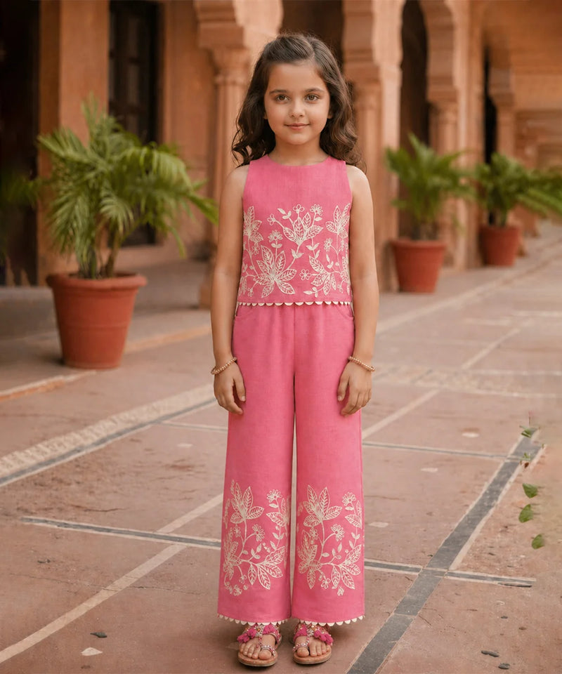 Young girl in a pink embroidered outfit standing in an outdoor setting with plants and architectural elements.