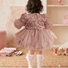 A toddler viewed from behind wearing a dusty pink ruffled tulle dress with puffed sleeves and a bow at the waist, paired with white lace socks and white shoes, standing in a soft pink nursery setting.