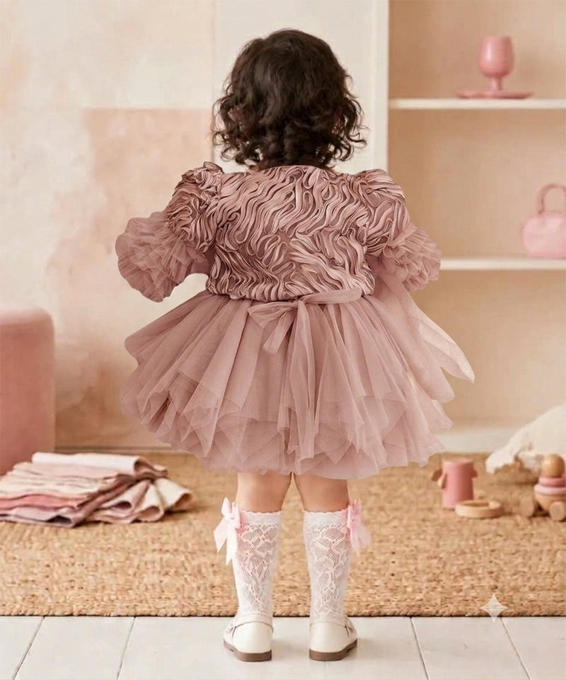 A toddler viewed from behind wearing a dusty pink ruffled tulle dress with puffed sleeves and a bow at the waist, paired with white lace socks and white shoes, standing in a soft pink nursery setting.