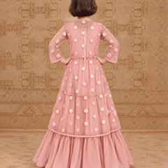A young girl viewed from behind wearing a dusty pink tiered gown decorated with white floral embroidery, standing against a patterned beige backdrop.