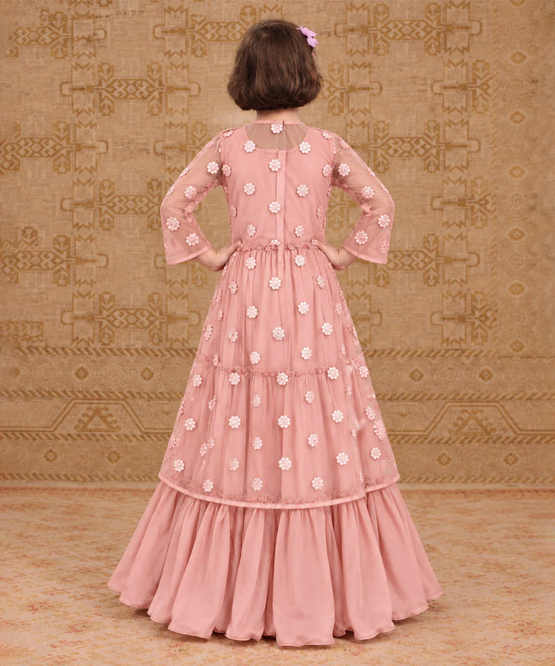 A young girl viewed from behind wearing a dusty pink tiered gown decorated with white floral embroidery, standing against a patterned beige backdrop.