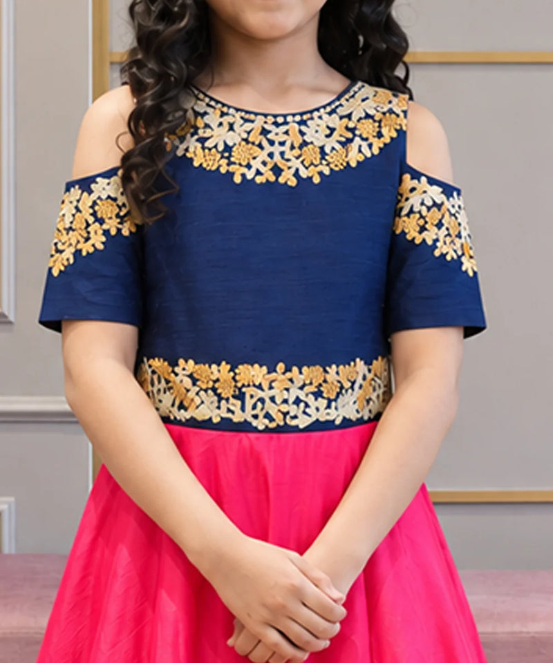 A child wearing a cold-shoulder navy blue top with gold and cream floral embroidery, paired with a bright pink skirt.