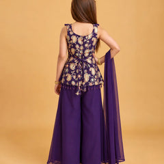 A woman shown from the back wearing a purple traditional outfit with a heavily embroidered peplum-style top, wide-leg pants, and a matching sheer dupatta, set against a solid warm beige studio background.