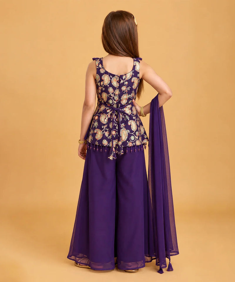 A woman shown from the back wearing a purple traditional outfit with a heavily embroidered peplum-style top, wide-leg pants, and a matching sheer dupatta, set against a solid warm beige studio background.