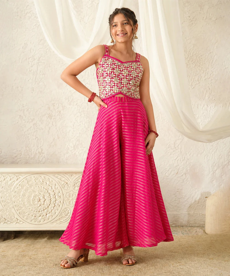 Woman wearing a pink and white patterned top with a long pink skirt against a white curtain background.