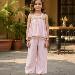 Young girl in a pink outfit standing outdoors with plants and furniture in the background