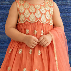 A child wearing a sleeveless peach-orange dress with a heavily embroidered gold leaf-patterned bodice and scattered gold floral motifs on the tulle skirt, paired with a matching sheer dupatta.