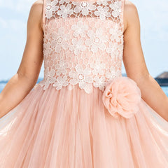 A sleeveless peach pink tulle dress with a white floral lace bodice, featuring a large matching peach pink fabric flower on the side, shown on a child model against a blue sky background.