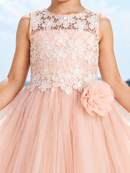 A sleeveless peach pink tulle dress with a white floral lace bodice, featuring a large matching peach pink fabric flower on the side, shown on a child model against a blue sky background.