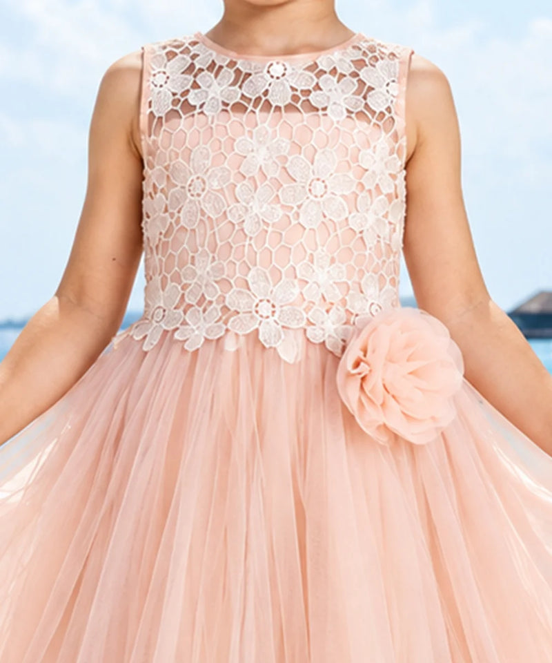 A sleeveless peach pink tulle dress with a white floral lace bodice, featuring a large matching peach pink fabric flower on the side, shown on a child model against a blue sky background.