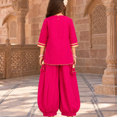 A woman viewed from behind wearing a bright pink traditional outfit with gold trim, consisting of a short-sleeve top and matching loose pants, standing in front of a historic stone building.