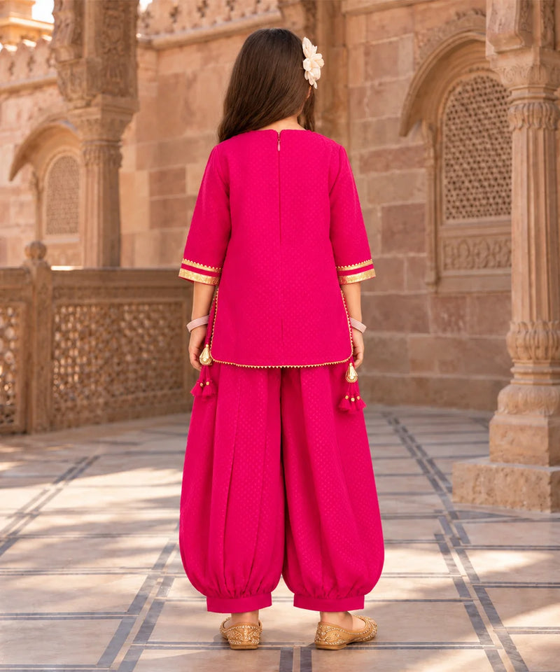 A woman viewed from behind wearing a bright pink traditional outfit with gold trim, consisting of a short-sleeve top and matching loose pants, standing in front of a historic stone building.