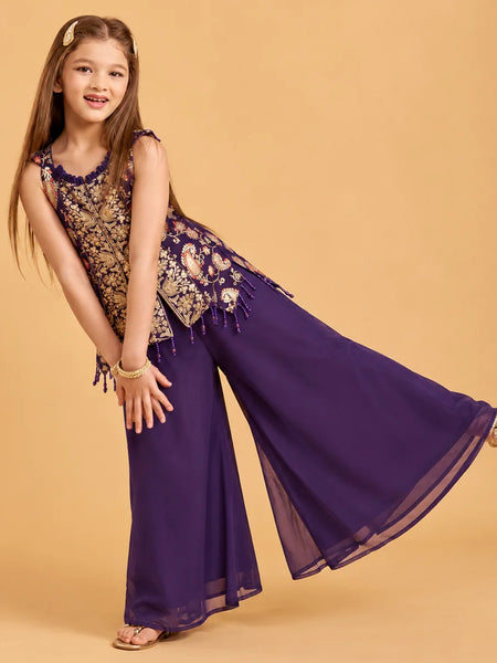 A young girl modeling a purple and gold embroidered traditional outfit with wide-leg pants, posed against a solid beige studio background.