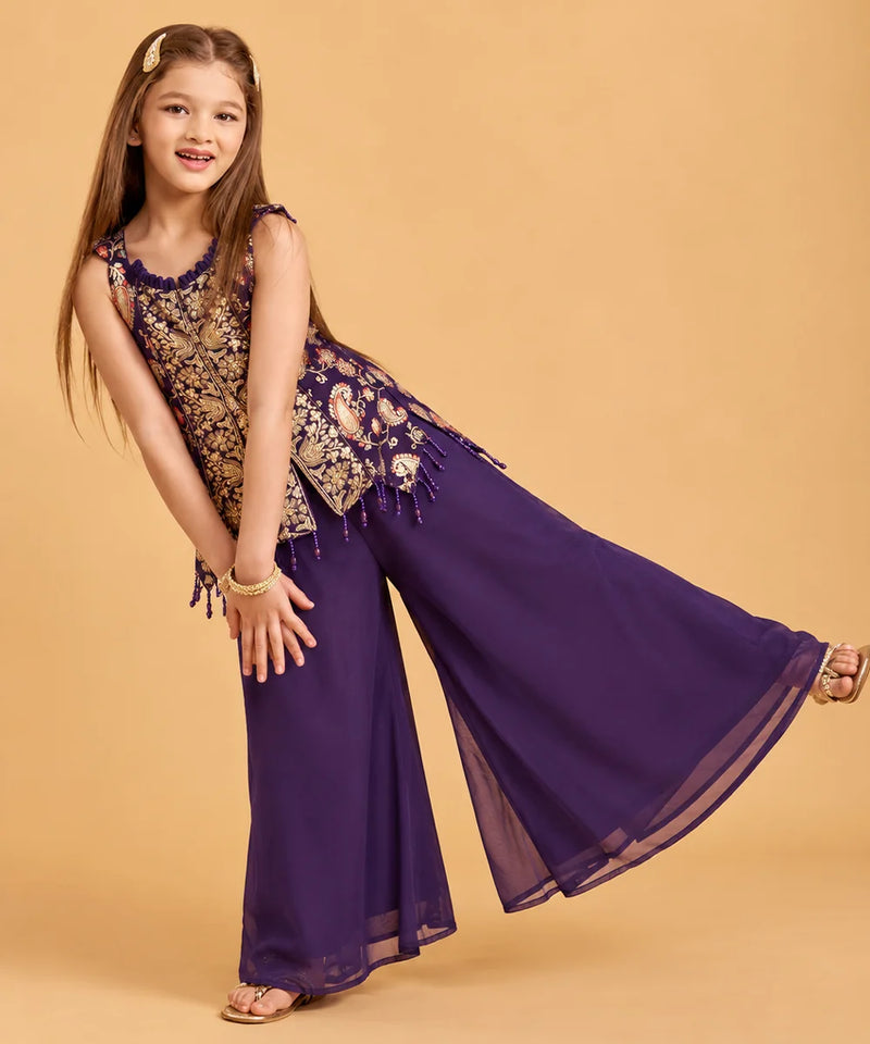 A young girl modeling a purple and gold embroidered traditional outfit with wide-leg pants, posed against a solid beige studio background.