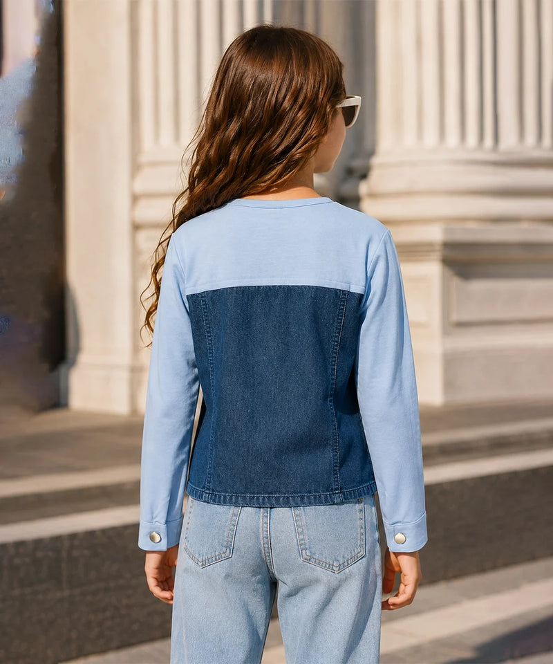 A child viewed from behind wearing a color-block denim jacket with light blue sleeves and upper back, and a darker blue denim back panel, paired with light-wash jeans in an outdoor setting with stone columns.