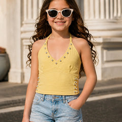 A young girl with long wavy brown hair wearing a yellow studded halter top and denim shorts, posing outdoors in front of a classical building with white columns.