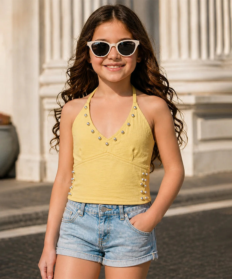 A young girl with long wavy brown hair wearing a yellow studded halter top and denim shorts, posing outdoors in front of a classical building with white columns.