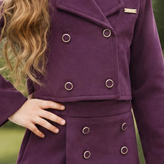 A close-up of a person wearing a double-breasted purple coat with silver-tone metal grommets and a small gold nameplate on the lapel, photographed outdoors against a blurred green background.