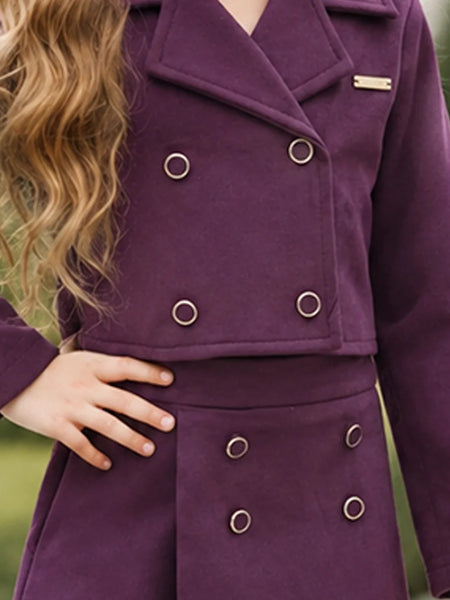 A close-up of a person wearing a double-breasted purple coat with silver-tone metal grommets and a small gold nameplate on the lapel, photographed outdoors against a blurred green background.