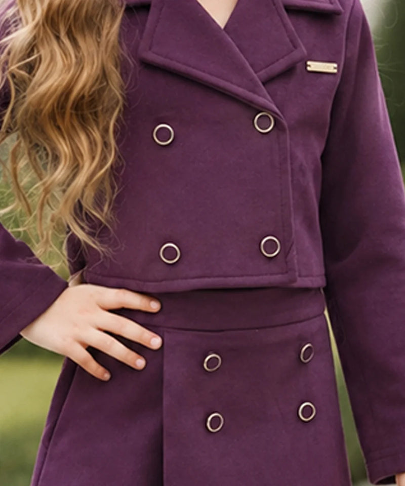 A close-up of a person wearing a double-breasted purple coat with silver-tone metal grommets and a small gold nameplate on the lapel, photographed outdoors against a blurred green background.