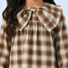 A close-up of a child wearing a brown and beige plaid long-sleeve top with a large fabric bow at the collar, set against a light blue background.