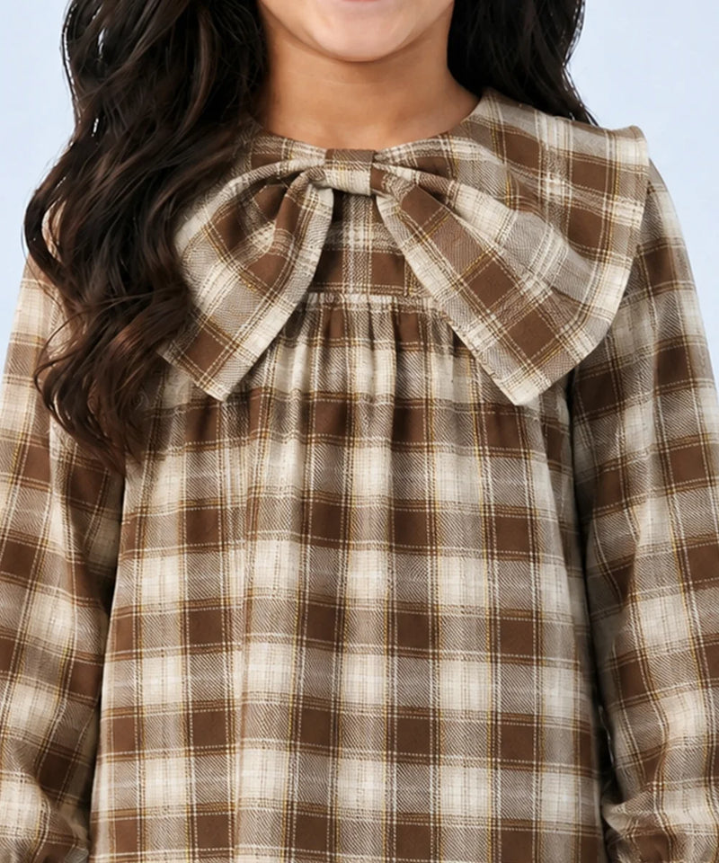 A close-up of a child wearing a brown and beige plaid long-sleeve top with a large fabric bow at the collar, set against a light blue background.