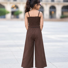 A child model shown from behind wearing a brown two-piece outfit consisting of a smocked camisole top and matching wide-leg trousers, standing in an outdoor plaza.
