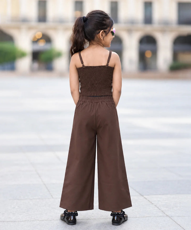 A child model shown from behind wearing a brown two-piece outfit consisting of a smocked camisole top and matching wide-leg trousers, standing in an outdoor plaza.